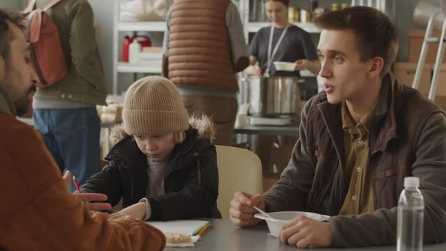 Medium slowmo of single father with little son and refugee man sitting at table in local food bank having lunch and talking about life