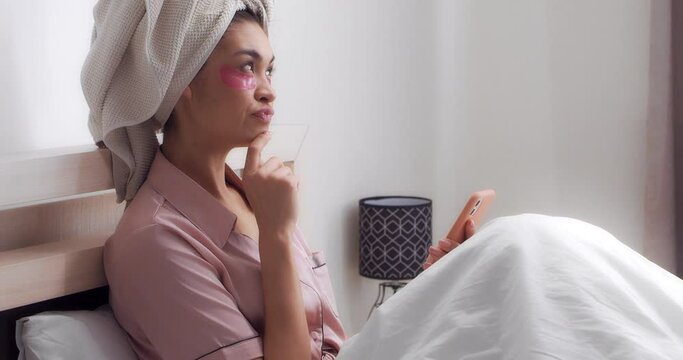Young woman in satin pink pajamas with a towel on head and patches on her eyes Is sitting under a white blanket on bed in bedroom, planning a bachelorette party,making a to-do list on her cell phone