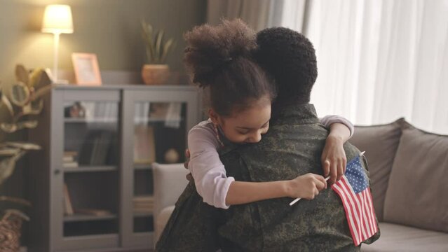 Medium slowmo of delighted African American little girl with US flag in hands running and embracing her military father returning home from long separation