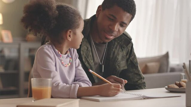 Waist up slowmo of African American military man helping his 6 year old daughter with homework, sitting at desk in living room