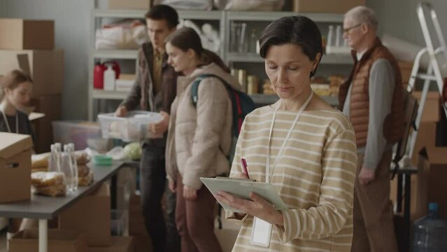 Medium slowmo of mature Caucasian female volunteer with clipboard smiling at camera while her colleague giving food and water provisions to people in need, working together at local food bank