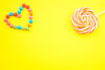 Lollipop and sweet multicolored chocolates laid out in the shape of a heart. Yellow background. Space for text.