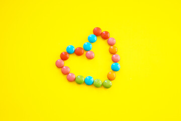 Sweet multi-colored chocolates, laid out in the shape of a heart. Yellow background. Space for text.