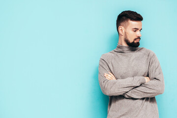 Handsome smiling model. Sexy stylish man dressed in grey turtleneck sweater and jeans. Fashion hipster male posing near blue wall in studio. Isolated. Crossed arms