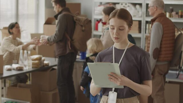 Medium slowmo portrait of young female volunteer with clipboard looking at camera while her colleague giving food and water supplies to refugees, working together at local food bank