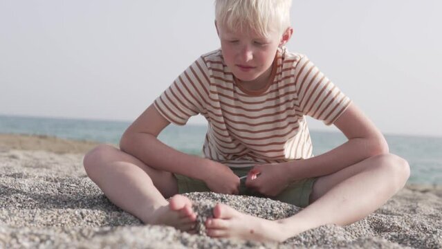 A beautiful blond boy sits on the beach by the sea and picks up the sand with his feet.