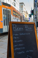 Advertisement of an italian restaurant, in Milan, Italy. Translation: "Naples Pizza; firsts; seconds; Starters; and many more". Blurred tram in the background.