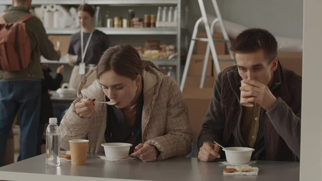 Medium slowmo of young Caucasian couple of refugees eating hot soup from disposable tableware sitting at table in local food bank with people standing in queue for their portion in background