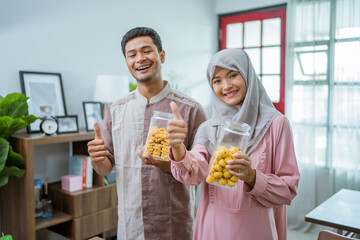 asian muslim man and woman with nastar snack looking at camera smiling. eid mubarak celebration