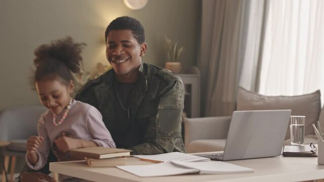 Medium slowmo of young Black man in military uniform working on laptop while staying at home together with playful 6 year old daughter