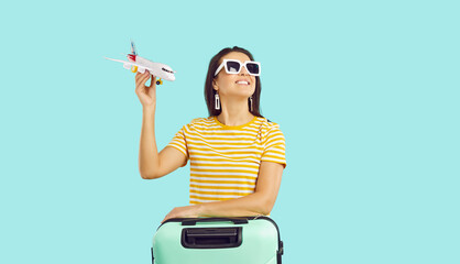 Happy woman traveler in sunglasses with suitcase hold airplane model in hands isolated on green...