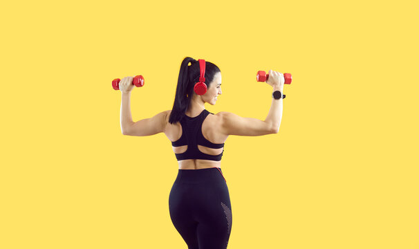 Backside shot of beautiful muscular Caucasian woman in sports crop top, leggings, wearable smart watch and red headphones isolated on yellow background listening to music and holding workout dumbbells