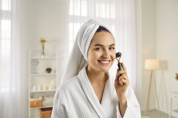 Portrait of pretty woman in bath towel and bathrobe massaging her face with trendy facial beauty...