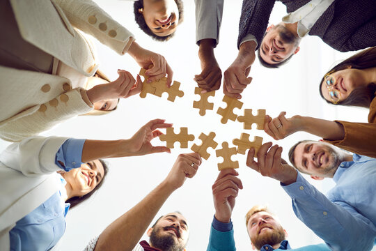 Teamwork. Happy different people put together pieces of wooden puzzles that symbolize teamwork. Smiling young multiracial people stand in circle and each holds one piece of puzzle. Bottom view.