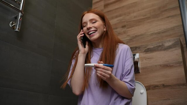 Cheerful young woman holding positive result on pregnancy test and talking by smartphone with husband sitting on toilet bowl. Happy excited lady telling boyfriend about pregnancy during phone call.