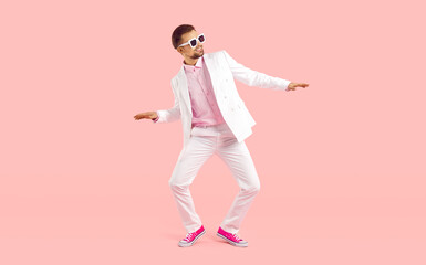 Cheerful cool young man in suit shows funny dance moves isolated on pink background. Funny trendy man in white formal suit, sneakers and glasses having fun. Full length body size view. Banner.