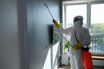 Worker in protective suit and face mask sprays chemicals indoors