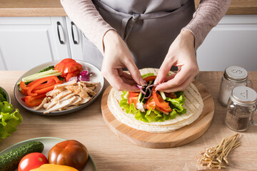 the hands of a young woman carefully lay out pieces of chicken fillet on green lettuce leaves on a grilled flatbread. preparation of buritto at home.