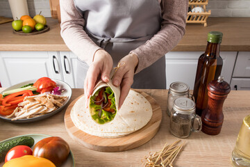 the hands of a young girl hold a wrapped buritto freshly cooked at home against the background of the kitchen. ingredients on the table.