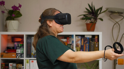 A girl in a green T-shirt wearing a virtual reality helmet stands in a room against a rack of games. In her hands are controllers. She is smiling. VR glasses for entertainment, learning and gaming.