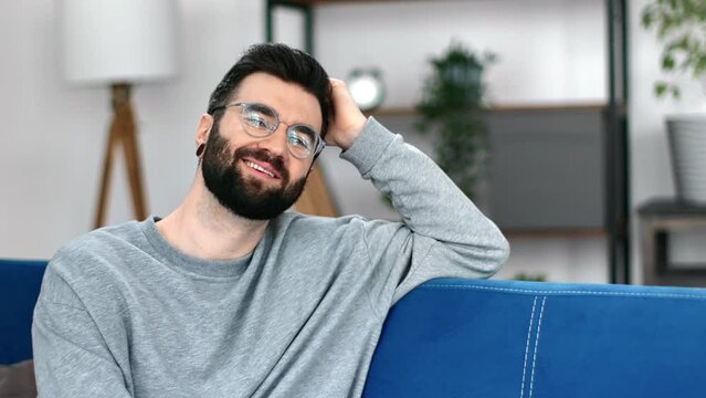Pleasant brunet modern hipster guy wearing eyeglasses and earrings sitting blue couch at apartment