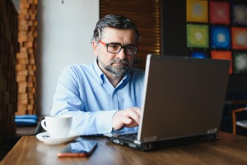 Mature businessman drinking coffee in cafe. Portrait of handsome man wearing stylish eyeglasses using laptop, looking at camera, smiling. Coffee break concept