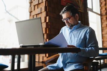 Mature businessman drinking coffee in cafe. Portrait of handsome man wearing stylish eyeglasses using laptop, looking at camera, smiling. Coffee break concept