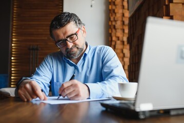 Obraz premium Mature businessman drinking coffee in cafe. Portrait of handsome man wearing stylish eyeglasses using laptop, looking at camera, smiling. Coffee break concept