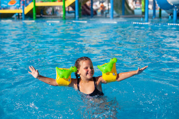 Naklejka premium Happy child is playing in pool. Little girl in inflatable armbands with dinosaurs is happy from swimming lessons in new water park. 