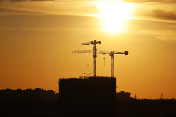 Silhouettes of two tower cranes and unfinished buildings at sunset. Housing construction, apartment block in city on dramatic sky background