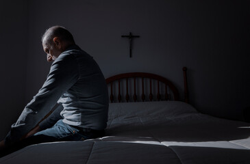 Side view of adult man sitting on bed with religious cross on wall