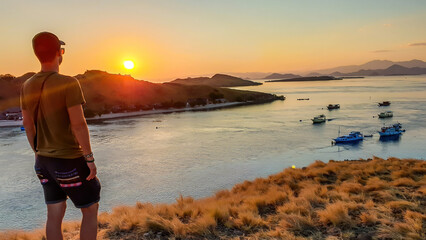 A man standing on top of a small island, facing a rising sun over Komodo National Park, Flores, Indonesia. Golden hour over the islands and sea. Some boats anchored to the bay. New day begining