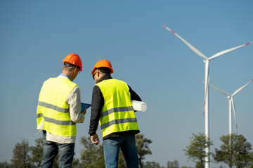 Wind turbines generate electric energy in front of engineers. Engineers in protective helmets stand holding project papers roll against rotating windmills