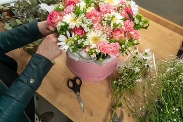 Close-up of the hands of a young female florist packing a beautiful composition of delicate pink roses, carnations and white daisies into a cardboard box and paper on the table.