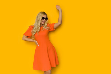 Happy blond woman in orange cocktail dress is looking to the side and waving hand.