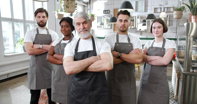 Diverse group of cafe employees in grey aprons stand next to each other, crossing hands with confidence. Staff of bright and cozy coffee bar looking into camera.