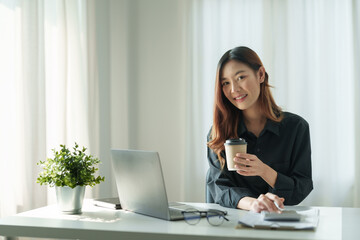 Charming asian female office worker working on a laptop computer and enjoying drinking coffee in a modern office.