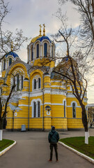 A young man in a thick jacket stands in front of St Volodymyr's Cathedral. The cathedral is painted yellow, with blue and golden-domed rooftop. Overcast and cold day.