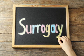Fototapeta premium Woman writing word Surrogacy on small blackboard at wooden table, top view