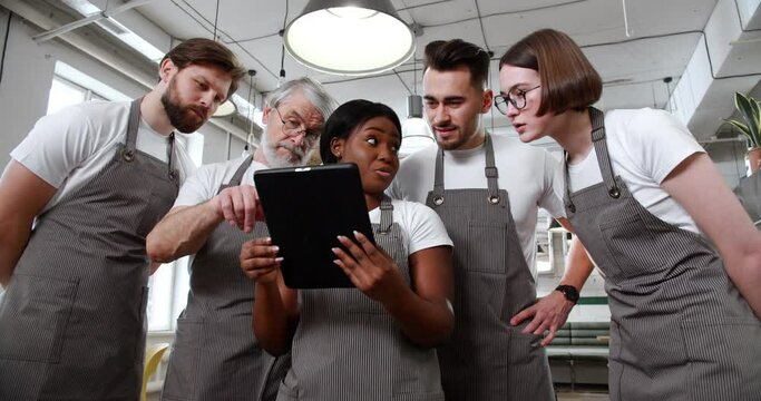 Young African American woman in grey apron gives presentation to colleagues using tablet, nodding and gesturing. Group of cafe employees having a meeting at workplace. - Powered by Adobe