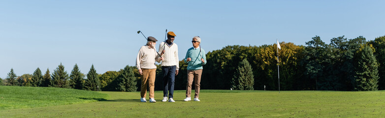 senior interracial friends walking with golf clubs on green field, banner.