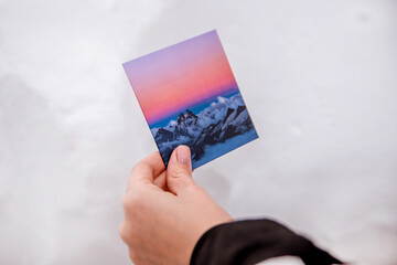 A hand holds a photo with mountains on a background of snow