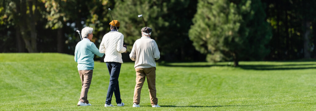 back view of senior multiethnic friends walking with golf clubs on green field near trees, banner.
