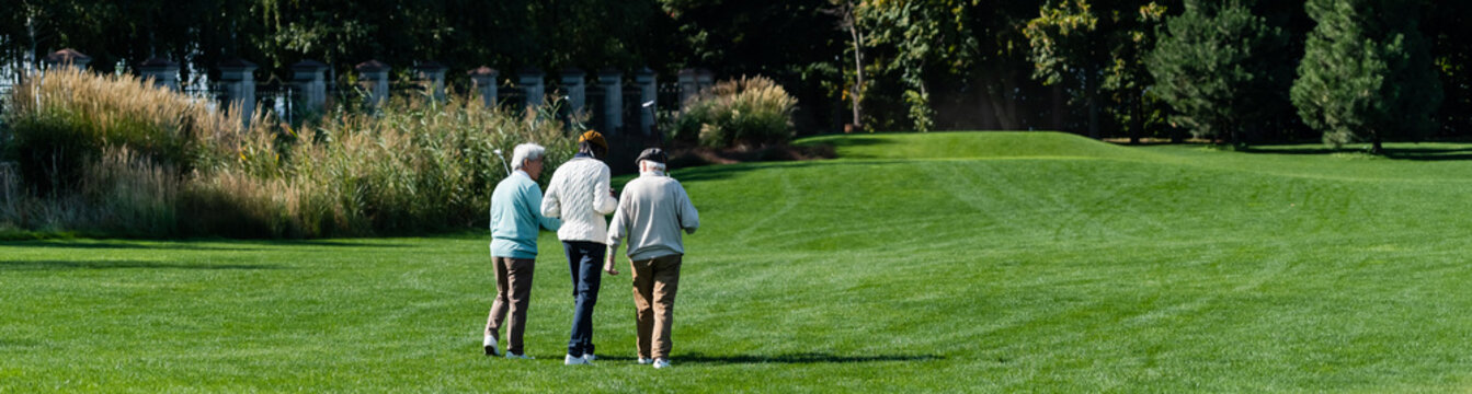 back view of senior interracial friends walking on green field with golf clubs, banner.