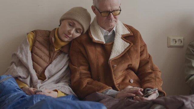 Slowmo of adult Caucasian couple of refugees in old warm clothes hiding with other people in shelter indoors. Woman napping on husbands shoulder while man trying to connect with family on mobile phone