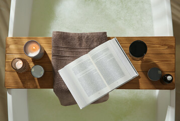 Wooden bath tray with open book, glass of wine and cosmetic products on tub indoors, top view