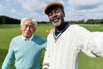 smiling african american man taking selfie with senior asian friend of golf field.