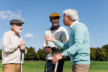 senior interracial friends holding golf clubs and talking on field.