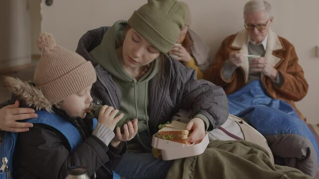 Slowmo of caring young Caucasian woman feeding her little son with sandwiches while staying in cold uncomfortable asylum with other refugees including mature couple sitting against wall in background