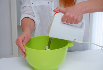 Home Сulinary Concept. Young woman baking a cake in the kitchen. Female using a handheld mixer to whisk the fresh ingredients in a green mixing bowl. Modern kitchen interior Cropped image.